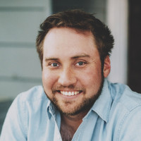 A man smiling casually, wearing a smart blue denim shirt. He’s sat in front of a house that’s blurred out behind him.